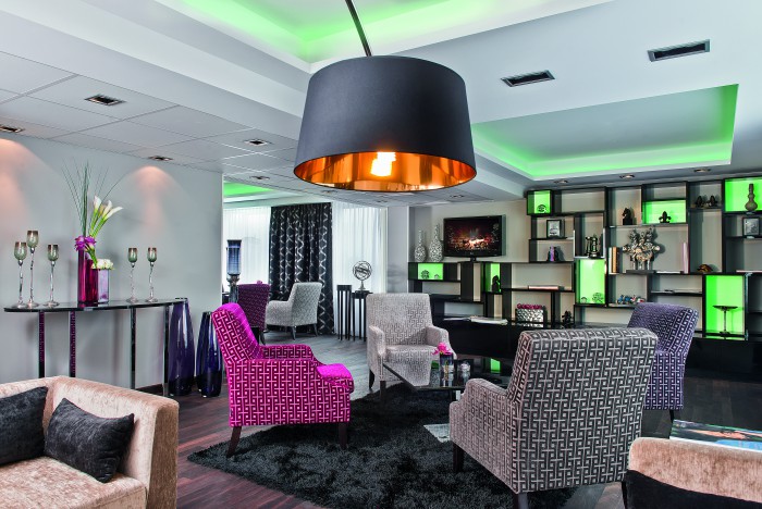Hotel Palace Moderne Club Lounge im Hotel Palace Berlin mit gemusterten Stühlen, schwarzem Teppich, großer Hängelampe und hinterleuchtetem Regal mit Dekorationsgegenständen. Ein Hauch von eleganter Raffinesse im Herzen Berlins.