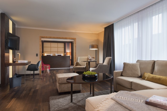 Die Senior Suite im Hotel Palace Berlin ist ein geräumiger Rückzugsort mit Holzböden und neutralen Farbtönen. Sie verfügt über einen Sitzbereich mit beigen Sofas, runden Couchtischen und einem großen Fenster mit transparenten Vorhängen.
