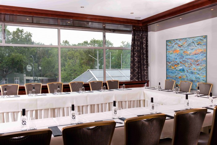 Tagungsraum im Hotel Palace Berlin mit großem U-förmigen Tisch, umhüllt von weißen Tischdecken und umgeben von Stühlen. Tageslicht strömt durch die Fenster mit Blick auf Bäume, während ein abstraktes Gemälde dem Raum Charakter verleiht.