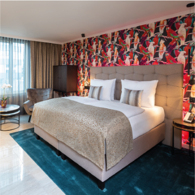 Business-Premium-Zimmer im Hotel Palace Berlin mit großem Bett, blumiger Tapete, Sessel, grauer Couch und Tageslicht durch transparente Vorhänge.