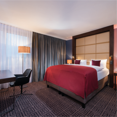Geräumiges Businesszimmer im Hotel Palace Berlin mit großem Bett, roter Bettwäsche und vier weichen Kissen. Ein Schreibtisch mit Stuhl und zwei Lampen ergänzt den eleganten Stil, während Vorhänge die Fenster für mehr Privatsphäre rahmen.