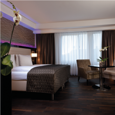 Modernes Clubzimmer im Hotel Palace Berlin mit Doppelbett, zwei Sesseln, kleinem Tisch und wandmontiertem Fernseher. Stilvolle Tapeten, dunkle Holzböden und eine dekorative Orchidee schaffen eine elegante Atmosphäre.