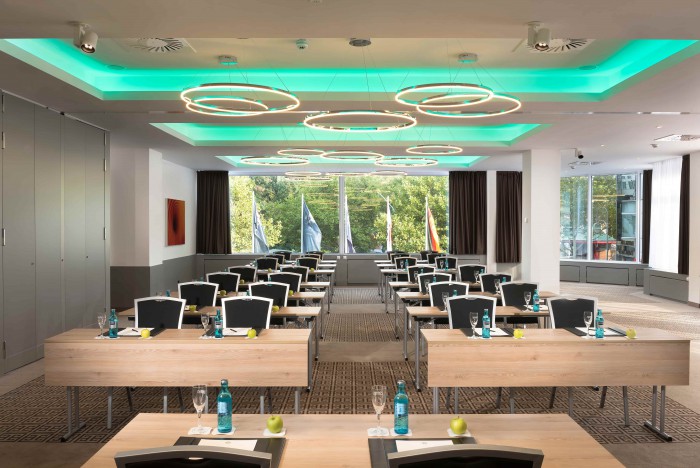 Moderner Tagungsraum im Hotel Palace Berlin mit Holztischen und schwarzen Stühlen in Reihen. Jeder Tisch ist mit Wasserflaschen, Notizblöcken und grünen Äpfeln ausgestattet. Runde Deckenleuchten und türkisfarbene Beleuchtung ergänzen die Ausstattung